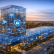 Illustration d’un bâtiment moderne représenté avec une surcouche numérique en transparence, mettant en évidence une infrastructure intelligente et sécurisée avec des connexions réseau, des icônes de sécurité et des composants technologiques.